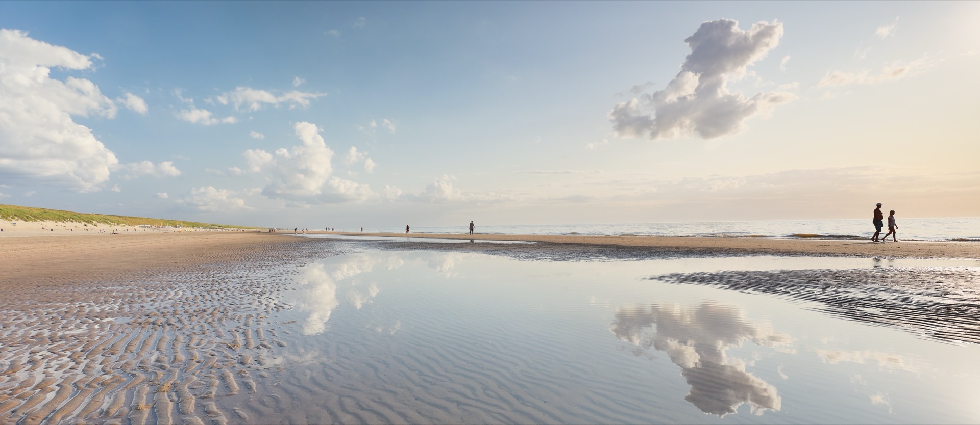 Laagwater op het strand van de Noordkop, met wolken die weerspiegelen in een geul en wandelaars aan de vloedlijn